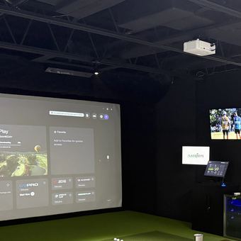 Both simulator bays at FlagShots in Dunedin, FL, seen from the lounge, with green turf hitting areas and a course projected on the impact screen