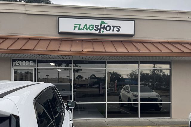 The FlagShots storefront in Dunedin, Florida: a white FlagShots lightbox sign above a copper awning and a glass frontage marked 2109 G, and free parking directly in front